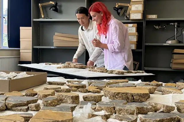 Un hombre que viste una camisa blanca y una mujer que viste una camisa morada están detrás de un escritorio con numerosos trozos coloridos de muralla romana en ellos.