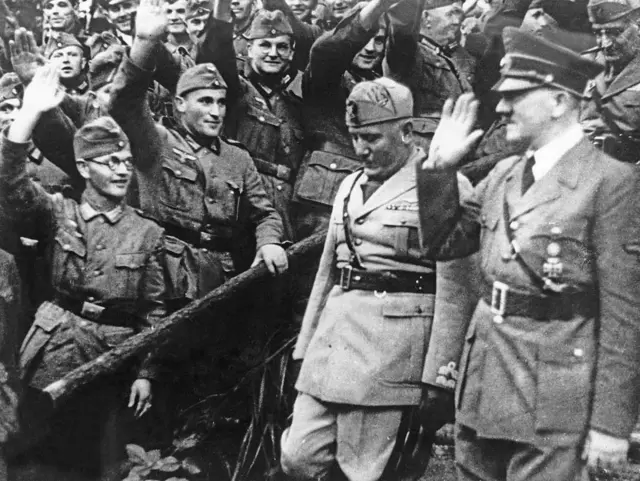 Hitler saluda a las tropas que lo aclaman haciendo el saludo fascista. A su lado Mussolini camina cabizbajo.