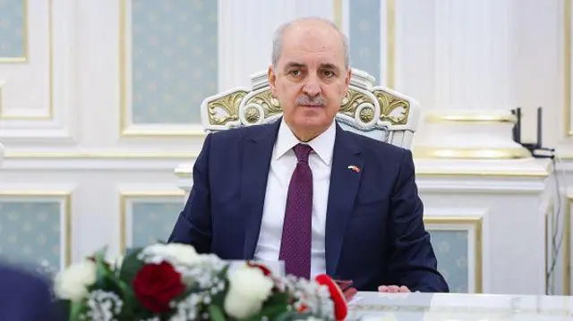 Numan Kurtulmuş bir sandalyede takım elbiseli, ön planda beyaz ve kırmızı güllerden oluşan bir çiçek süslemesi görünüyor.
