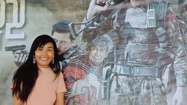 စစ်တပ်ဝါဒဖြန့်ဇာတ်ကားတစ်ခုရဲ့ပိုစတာရှေ့မှာ မြင်ရတဲ့ အဆိုတော် လီလီနိုင်ကျော် 