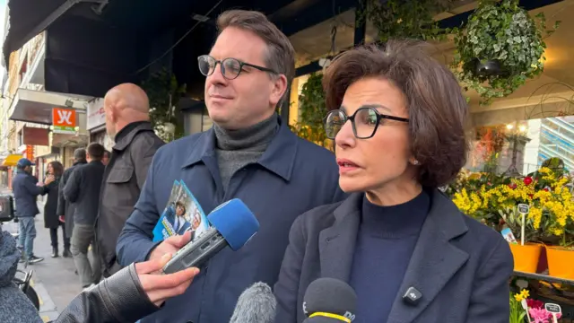 Kobieta w czarnych okularach mówi do mikrofonu. Za nią, po lewej, stoi ubrany w granatowy płaszcz członek kampanii 