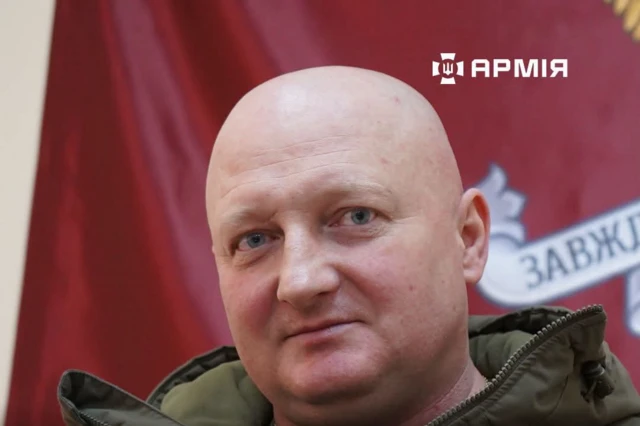 Портрет крупним планом чоловіка середнього віку на тлі червоного прапора. Людина одягнена в темно-оливкову куртку. На задньому плані частково видно емблему та фрагмент напису на стрічці. Фото добре освітлене, основна увага зосереджена на обличчі в центрі кадру.