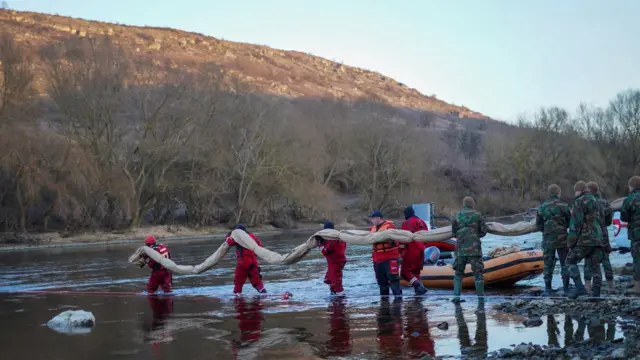 Группа спасателей и людей в камуфляже стоит в неглубокой воде реки. Они несут длинный гибкий барьер, вероятно предназначенный для сдерживания загрязнения. На берегу рядом видно надувную лодку. На заднем плане — холм и деревья без листьев.