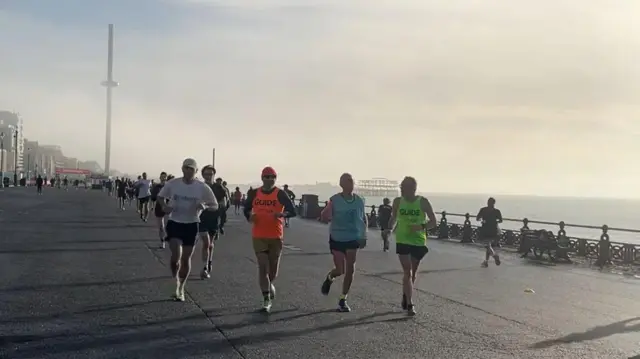Keith corre en fila con sus dos guías. Está atado a uno de ellos, Terry Westley, quien le da instrucciones verbales y físicas. Corren por el paseo marítimo de Hove Lawns, junto a la playa de Hove. El cielo se ve brumoso y brillante, con la Brighton i360, el Brighton Palace Pier y el Brighton West Pier visibles en el horizonte. Los corredores que completan el Hove Promenade Parkrun corren el recorrido detrás de ellos.