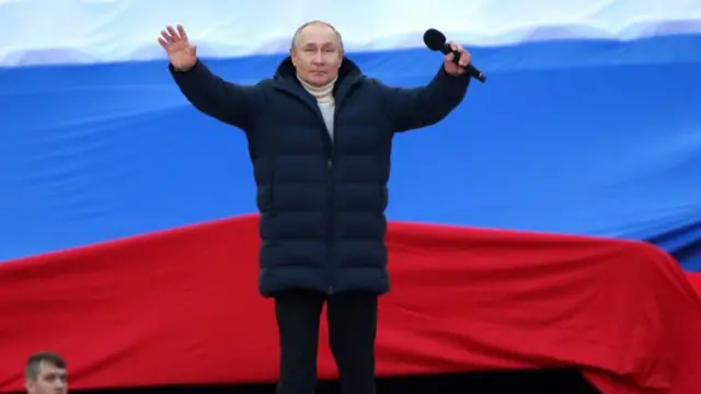 Putin, alza sus brazos de pie en un escenario con la bandera rusa detrás. 