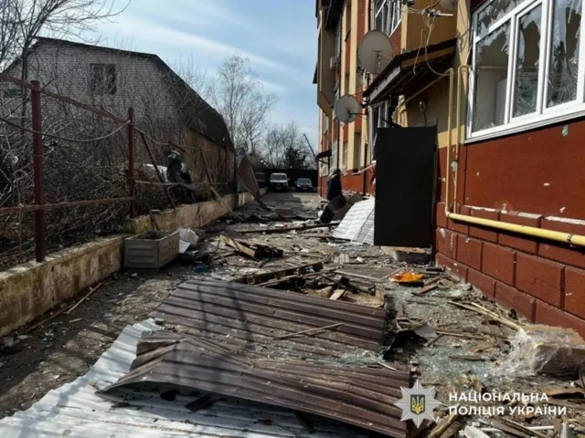 Видно нижнюю часть пострадавшего жилого дома. Вход в подъезд открыт, ближайшее окно выбито, внизу валяются обломки и осколки стекла.