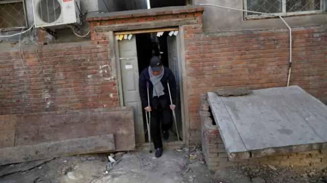 Hombre sale en muletas de una casa semiderruida de ladrillos rojos.