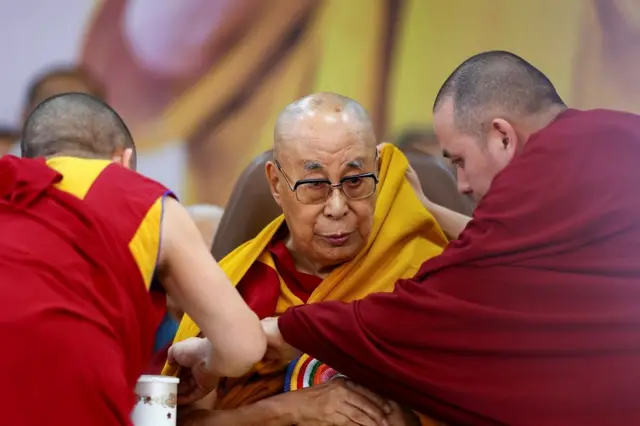 धर्मशालामा आफ्नो ९० औँ जन्मोत्सवमा सहभागी हुँदै दलाई लामा। रातो रङ्गको पोशाक लगाएका दुई जना धर्म गुरुले पहेलो गम्छा उनलाई लगाइदिइरहेका छन्