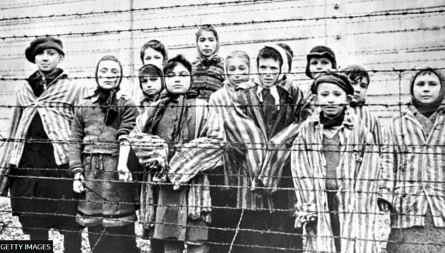 گروهی از کودکان بازمانده پشت یک حصار سیم خاردار در اردوگاه کار اجباری نازی‌ها در آشویتس