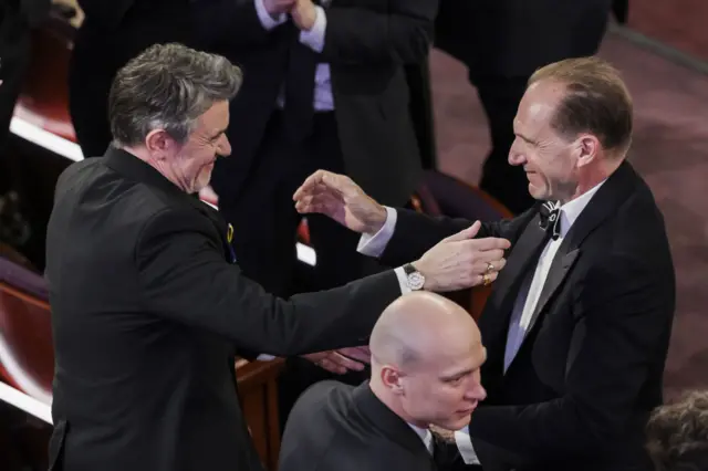 Dva muškarca u crnom smokinzima se smeju jedan drugom i gestikuliraju rukama kao da će da se zagrle na dodeli Oskara