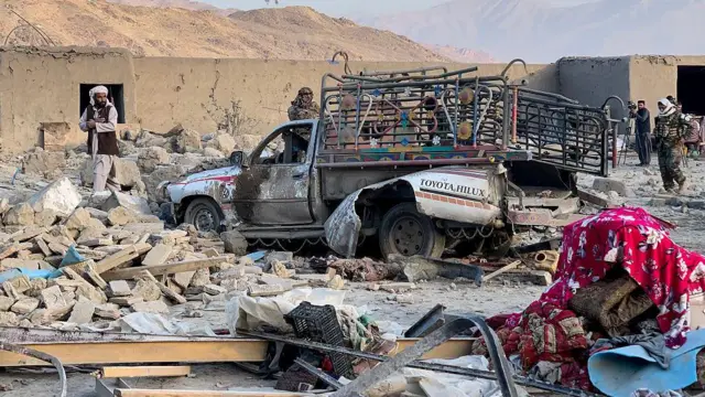 تصویر یک موتر تخریب شده در میان آوار. چند مرد نیز در محل حضور دارند. یک لحاف رنگی و تشک نیز در گوشه پایین سمت راست تصویر دیده می‌شود. 