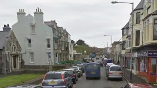 Calle Church en Port, con mucho tráfico. Tiene tiendas a un lado, una iglesia y casas adosadas al otro, con coches aparcados a ambos lados de la calle.