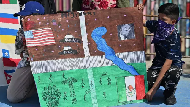 Dos niños migrantes centroamericanos, uno con gorra y el otro con la cara medio cubierta por un paliacate, sostienen una pancarta con un dibujo que muestra el muro fronterizo entre Estados Unidos y México, migrantes a un lado, agentes al otro, las banderas de ambos países y la cara del presidente estadounidense Donald Trump durante una protesta para exigir que se garanticen los derechos de migrantes y refugiados en el Día Mundial del Refugiado en Tijuana, Baja California, México, el viernes 20 de junio de 2025. (Foto de Carlos Moreno/Anadolu a través de Getty Images)