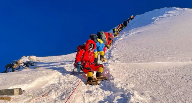 Bir grup yüzü maskeli ve oksijen aldıkları anlaşılan dağcı sıra halinde zirveden aşağıya iniyor. Hava günlük güneşlik yerde buzlu kar var.