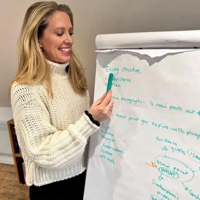 A woman with blonde hair wearing a cream jumper using a green pen on a large canvas of paper. 