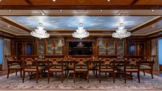 Amadea'nın yemek salonunda halı kaplı bir zemin üzerinde, büyük, koyu renkli ahşap bir masanın yanında ahşap sandalyeler var. Tavandan üç beyaz avize sarkıyor.