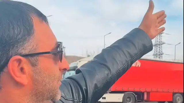 Водитель такси Абу, мужчина в черных очках и черной кожаной куртке, показывает рукой в сторону. На заднем плане стоит красная фура.