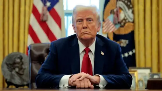 Perezida w'Amerika Donald Trump yicaye ku ntebe y'umusego areba muri 'camera' ashyize ibiganza ku meza, kimwe kigeretse ku kindi. Yambaye ikositimu y'ubururu, ishati y'umweru na karuvati itukura. Inyuma ye hari ibendera ry'Amerika. 