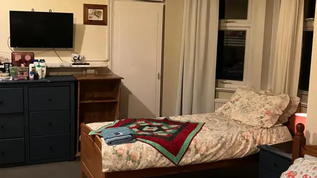 Una cama individual con ropa de cama de flores, una colcha de cuadros rojos y verdes y una toalla doblada. Hay un televisor en la pared, sobre dos muebles de madera. Hay artículos de aseo encima del armario.