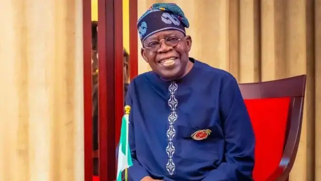 Onyeisiala Naịjirịa bụ Bola Tinubu yi uwe n'acha anụnụ anụnụ ma nọọrọ na ọche nwere akwa uhie uhie nakwa aja aja. Ọkọlọtọ Naịjirịa nọ ya n'ihu dịka ọ na amụrị ọnụ ọchi. O yi okpu n'acha anụnụ anụnụ ma yirikwa igobe anya.