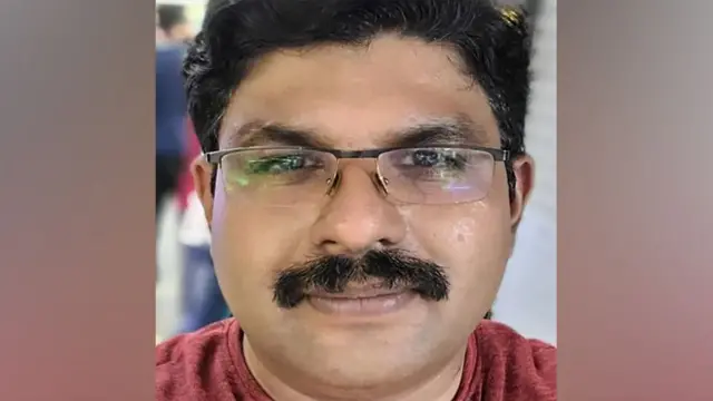 A close-up photograph of Deepak U facing the camera. He has short, dark hair, wears rectangular glasses, and has a thick moustache. His expression is neutral. He is wearing a dark red, short-sleeved T-shirt. The photograph appears to have been taken indoors. Behind him, the background is blurred, with indistinct figures of people and what look like closed shop shutters or metal doors, suggesting a public place