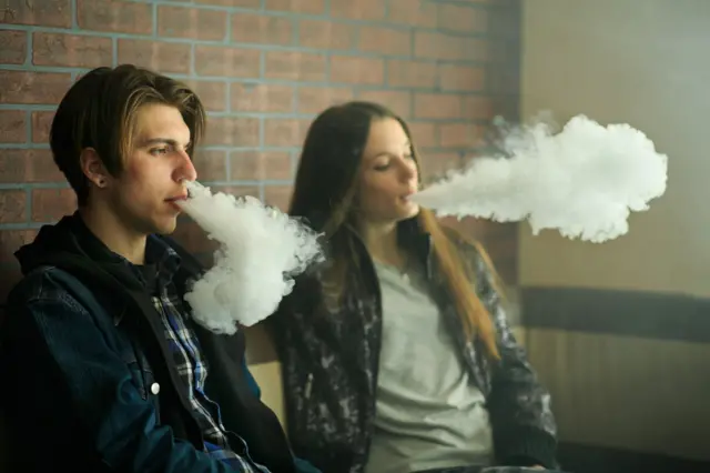 Two teenagers - a boy with floppy brown hair, wearing a checked shirt and navy coat, and a girl with long dark hair, wearing a grey top and patterned jacket, blow smoke from their mouths and nostrils.