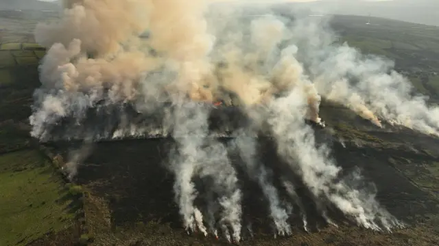 An aerial image of the wildfire at Corrick Road outside Draperstown. Plumes of grey smoke are spread across a large area of hillside. Much of the grassland is blackened but there is a green field to the left and green fields in the distance.