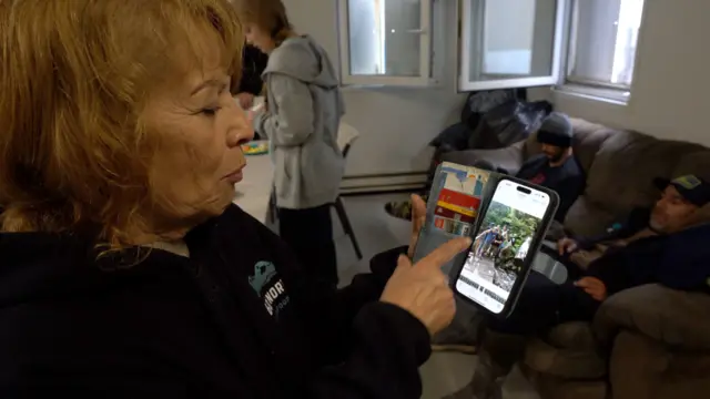 Uma senhorazebet ukpele e cabelos claros segura o celular e mostra uma foto para a câmera. No fundo, dois homens sentadoszebet ukum sofá. 