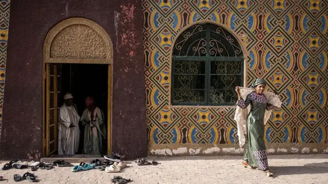 A woman, wearing a pink and green dress, arranges her veil. She walks in front of a patterned building.