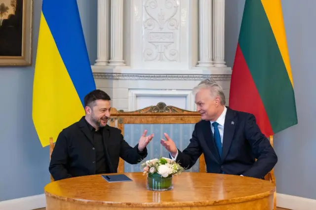 Зеленський і Науседа сидять за круглим дерев’яним столом у світлій залі з класичним інтер’єром. Вони активно жестикулюють, ніби обговорюють якесь питання в робочій, але невимушеній атмосфері. На столі стоїть невелика скляна ваза з композицією зі світлих квітів, поруч лежить темний блокнот або планшет. Позаду них розташовані великі прапори України та Литви.