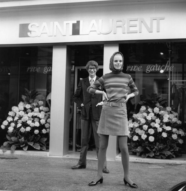O estilista francês Yves St Laurent do lado de fora de sua boutique Rive Gauche na França, com uma modelo vestindo uma saia e um suéter de sua coleção prêt-à-porter