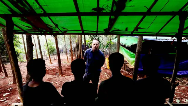Four young men stand with their backs to the camera, in a jungle under a tarpaulin roof. BBC correspondent Quentin Sommerville stands facing towards them.