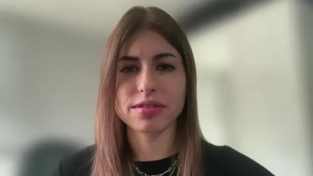 Sara tiene el pelo largo y castaño y una blusa negra. Mira directamente a la cámara.