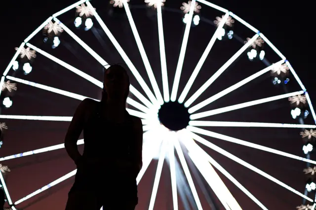 Silhueta de uma pessoa em primeiro plano, de pé e com as mãos atrás do corpo, diante de uma grande roda-gigante iluminada à noite. As luzes brancas da roda formam linhas radiais brilhantes ao fundo, criando um efeito visual circular e contrastando com o corpo escuro da pessoa.