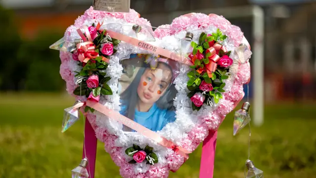 Un arreglo floral con forma de corazón. En el centro hay una foto de Chloe. Está ubicado en un parque.