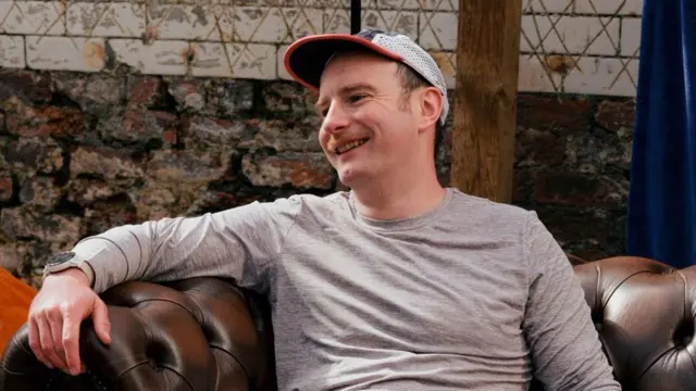 Xander, 37-letni mężczyzna, siedzi na sofie w szortach i koszulce do biegania. Śmieje się, a na nodze ma błoto.