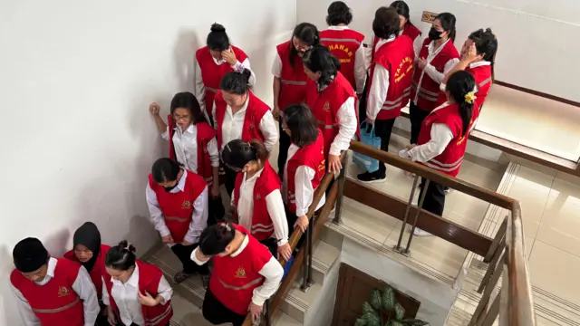 Belasan orang dengan baju kemeja putih dan rompi tahanan berwarna merah berdiri di tangga.