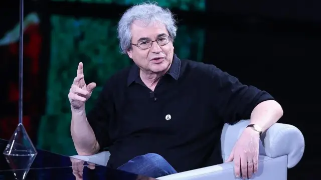 Rovelli, vestido com uma camisa preta e jeans, está sentado em um sofá cinza em um estúdio de televisão.