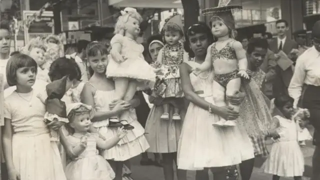 Foto em preto e branco mostra meninas brancas e negras de vestido segurando bonecas antigas