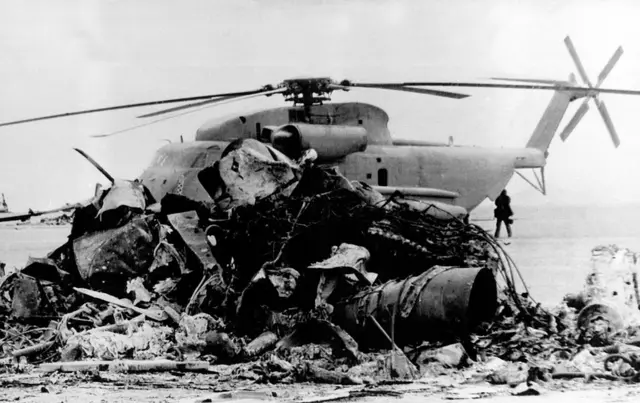 Купа обгорілих і сильно пошкоджених металевих уламків, серед яких можна розгледіти частини корпусу та двигуна. Це виглядає як залишки літака або вертольота після аварії чи вибуху. Метал чорний, деформований, з гострими краями, що свідчить про сильну пожежу.