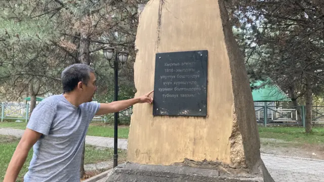 Элери Битикчи Үркүнгө арналган эстеликтеги азыркы жазууну көрсөтүп жатат