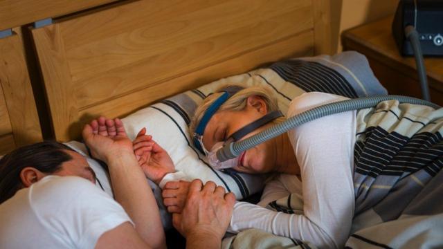 Mulher dormindo usando máscara de CPAP segurando a mão do marido em foto de banco de imagem