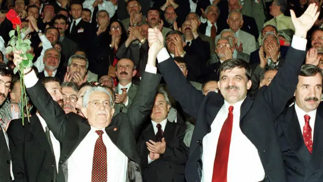Recai Kutan ve Abdullah Gül birlikte ellerini havaya kaldırıyor. Arkada kalabalıklar bir erkek grubu var. Herkes takım elbiseli. 