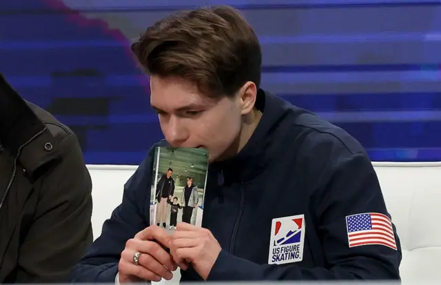 Максим Наумов в форме сборной США задумчиво сидит за столом, держа в руках фотографию своих родителей