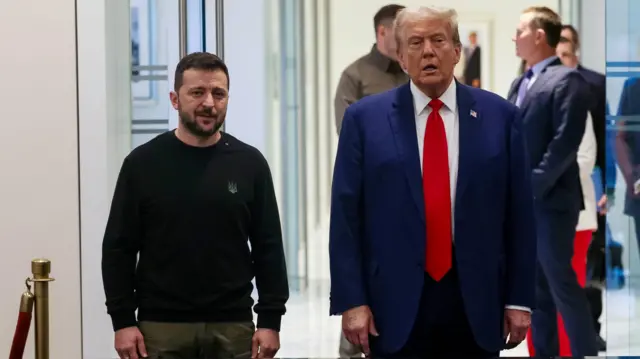 Volodymyr Zelensky and Donald Trump stand next to each other. Zelensky wears a black jumper and khaki trousers, while Trump wears a blue suit, white shirt and red tie. වොලොදිමීර් සෙලෙන්ස්කි සහ ඩොනල්ඩ් ට්‍ර්ම්ප් එකිනෙකා අසල සිටගෙන සිටියි. සෙලෙන්ස්කි කළු ටීෂර්ට් එකක් සහ කාකි කලිසමක් ඇඳ සිටින අතර ට්‍රම්ප් නිල් පැහැති ඇඳුමකින්, සුදු කමිසයකින් සහ රතු ටයි පටියකින් සැරසී සිටී.