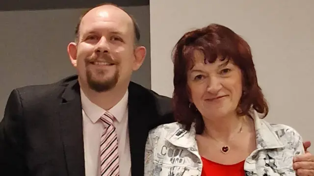 James Burley, que tiene cabello castaño, barba de chivo marrón y viste un traje negro, camisa blanca y corbata de rayas rosas, abraza a Natalie Zolty, que tiene cabello rojizo de longitud media, viste una chaqueta blanca con estampado negro, una blusa roja y un collar en forma de corazón.