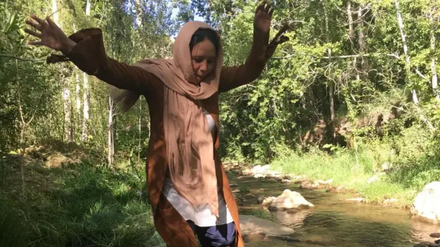 Zahra as an older girl wearing a headscarf and other flowing clothes, dancing along stones in a river. There are trees on both sides and the sun is shining through.