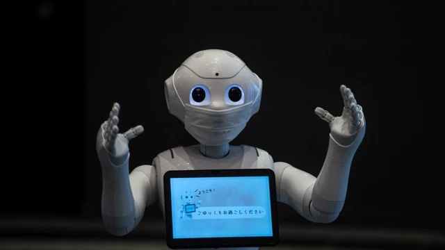 Um robô recepcionista, com os dois braços à frente, máscara no rosto e uma tela no peito.
