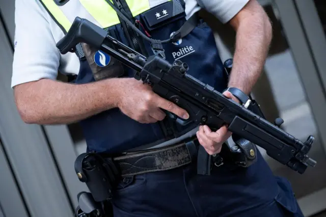 Agente de policía con chaleco y uniforme sosteniendo un arma larga frente a un edificio