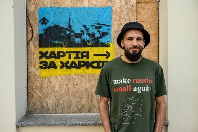Чоловік стоїть перед фанерою, на якій нанесений трафарет у жовто‑блакитних кольорах із зображенням міста та дронів і написом "Хартія → за Харків". Вона одягнена в темно‑зелену футболку з написом англійською "make russia small again" та силуетом карти.
